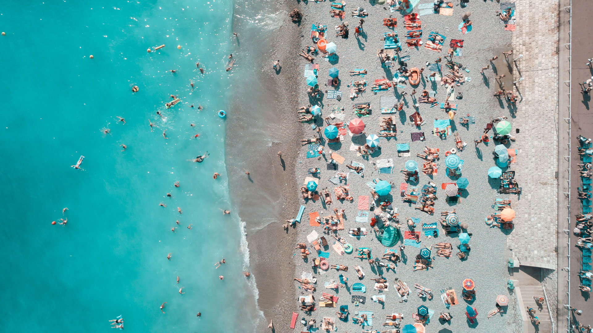 Aerial view of a bustling beach in Nice, France, with colorful umbrellas and sunbathers covering the pebbled shore, while swimmers and floaters enjoy the turquoise Mediterranean waters.