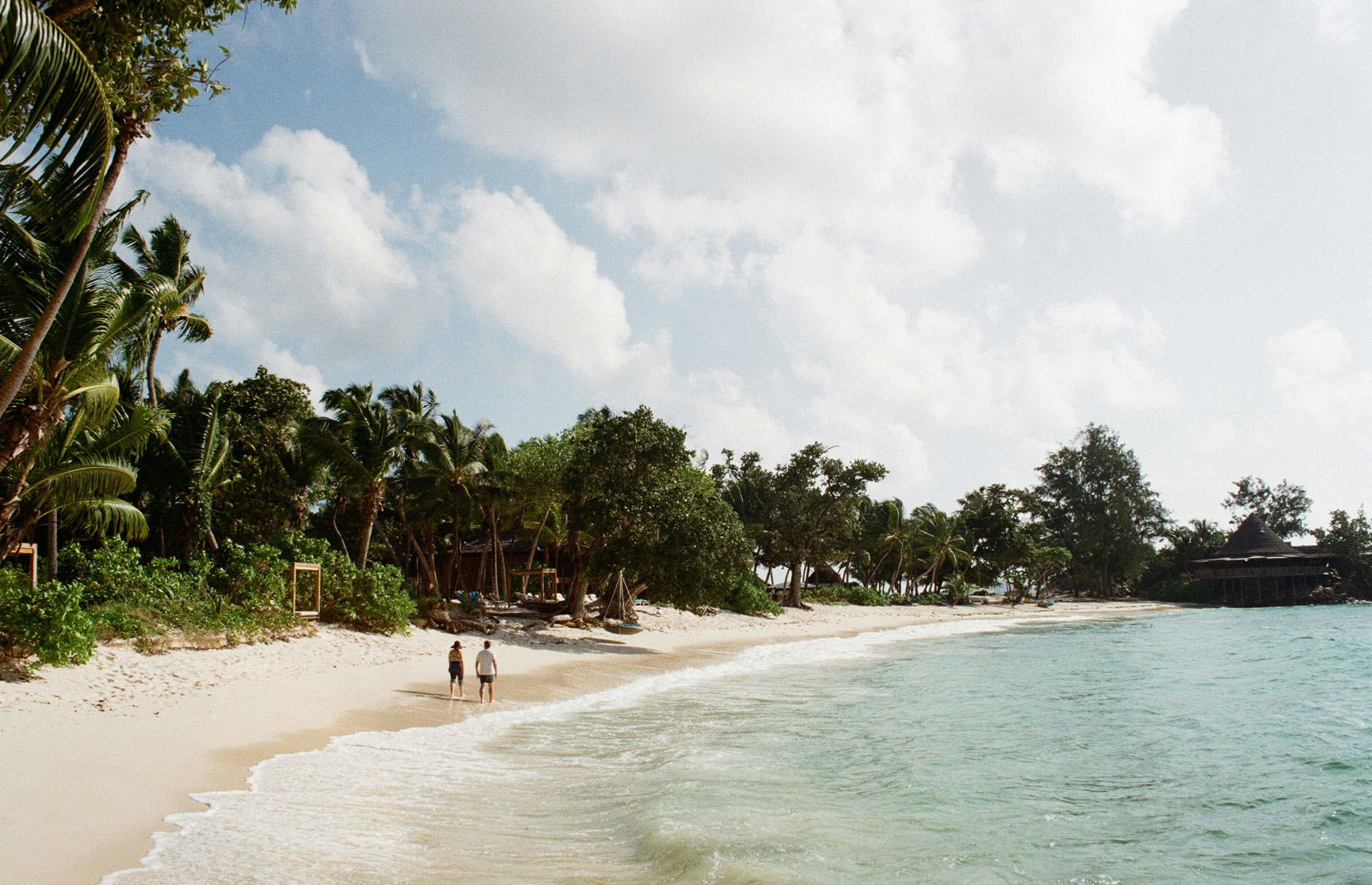 A serene beach scene in the Seychelles with two people walking along the soft white sand, bordered by lush tropical palm trees and gentle turquoise waves under a partly cloudy sky.