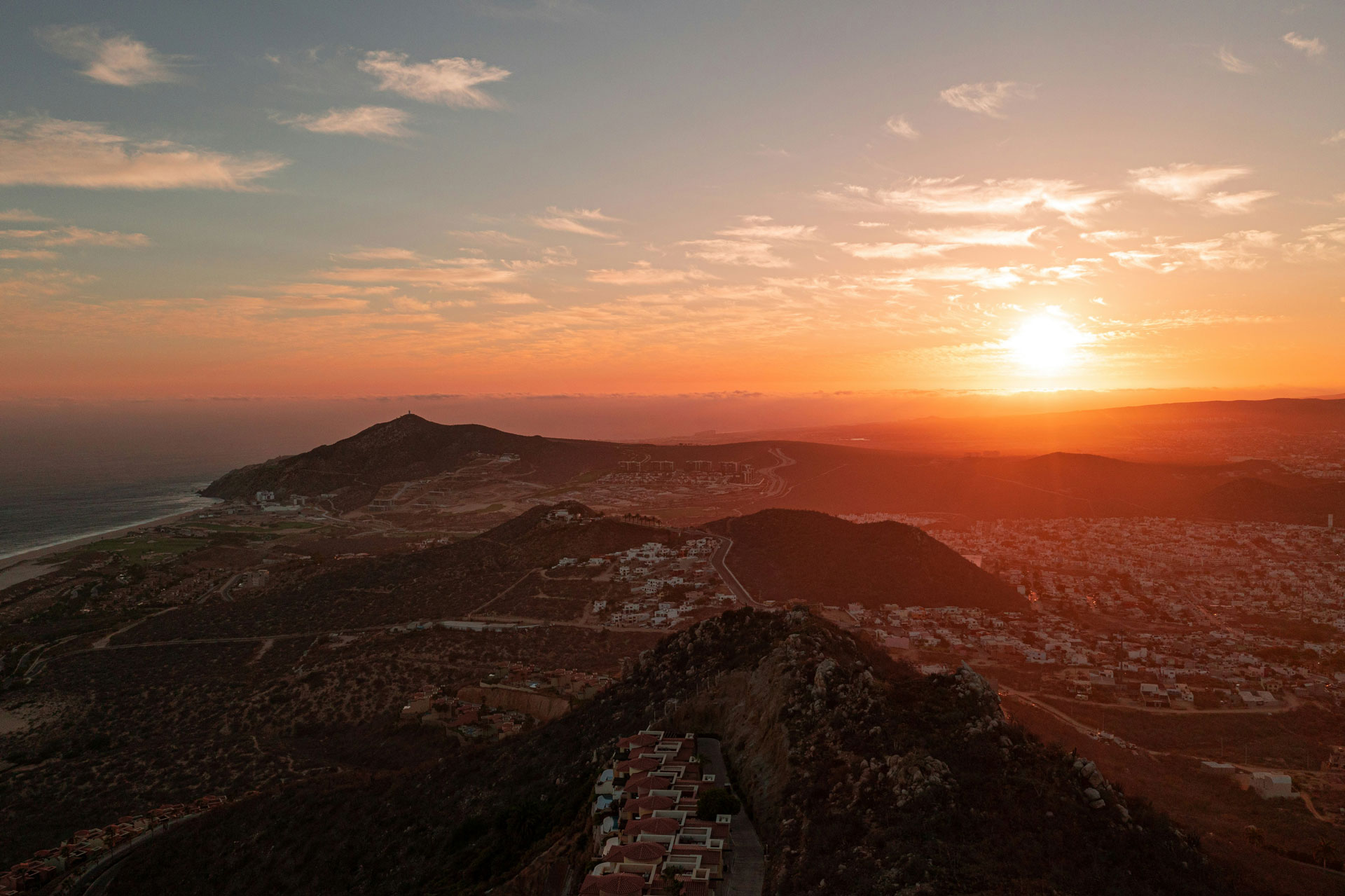 A breathtaking sunset over the rugged hills and coastal town of Los Cabos, Mexico, with the golden sun casting warm light across the landscape, silhouetting mountains, and highlighting the rooftops and winding roads below.