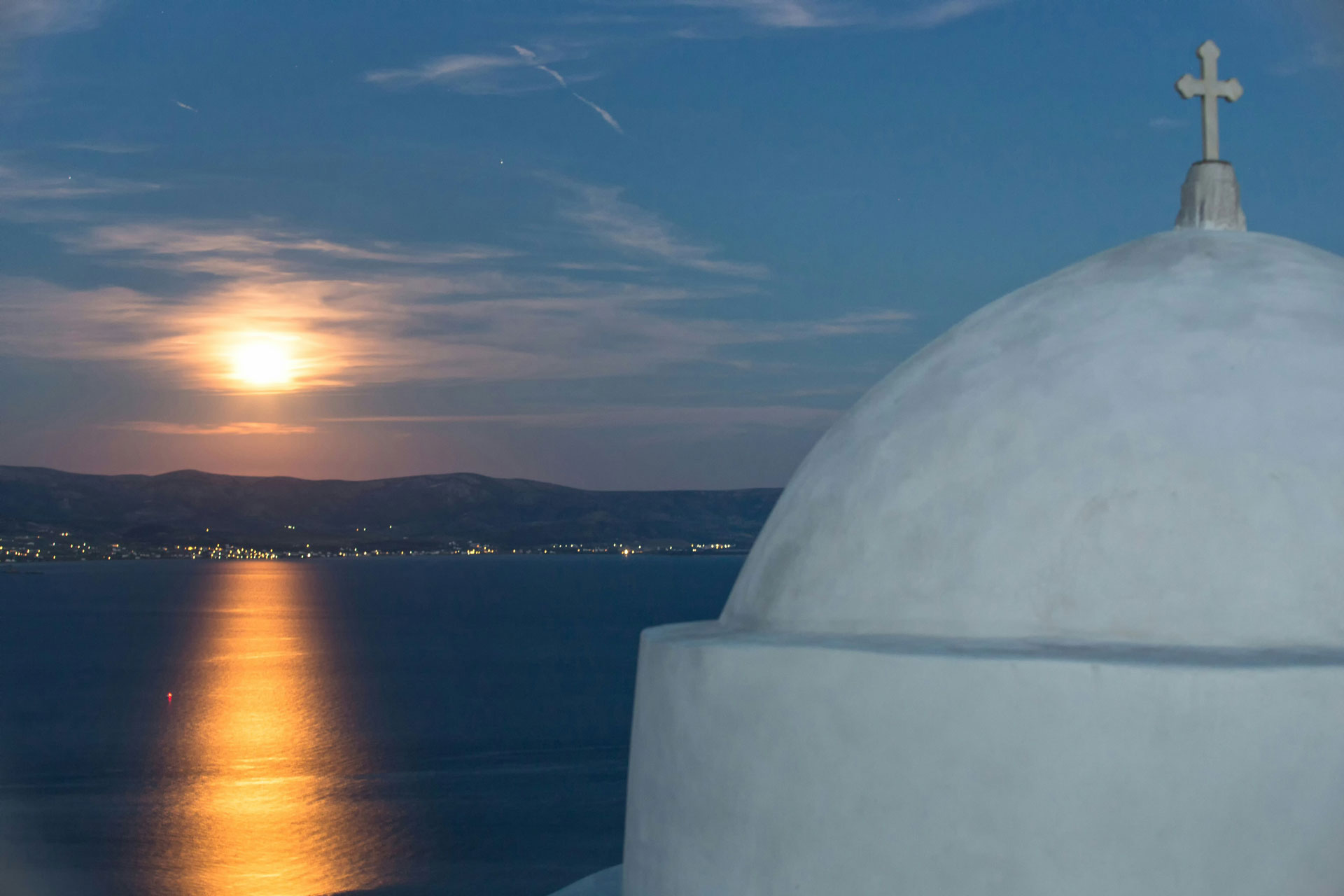 Nighttime view over the Aegean Sea from Paros, Greece, with a full moon casting a golden reflection on the water.