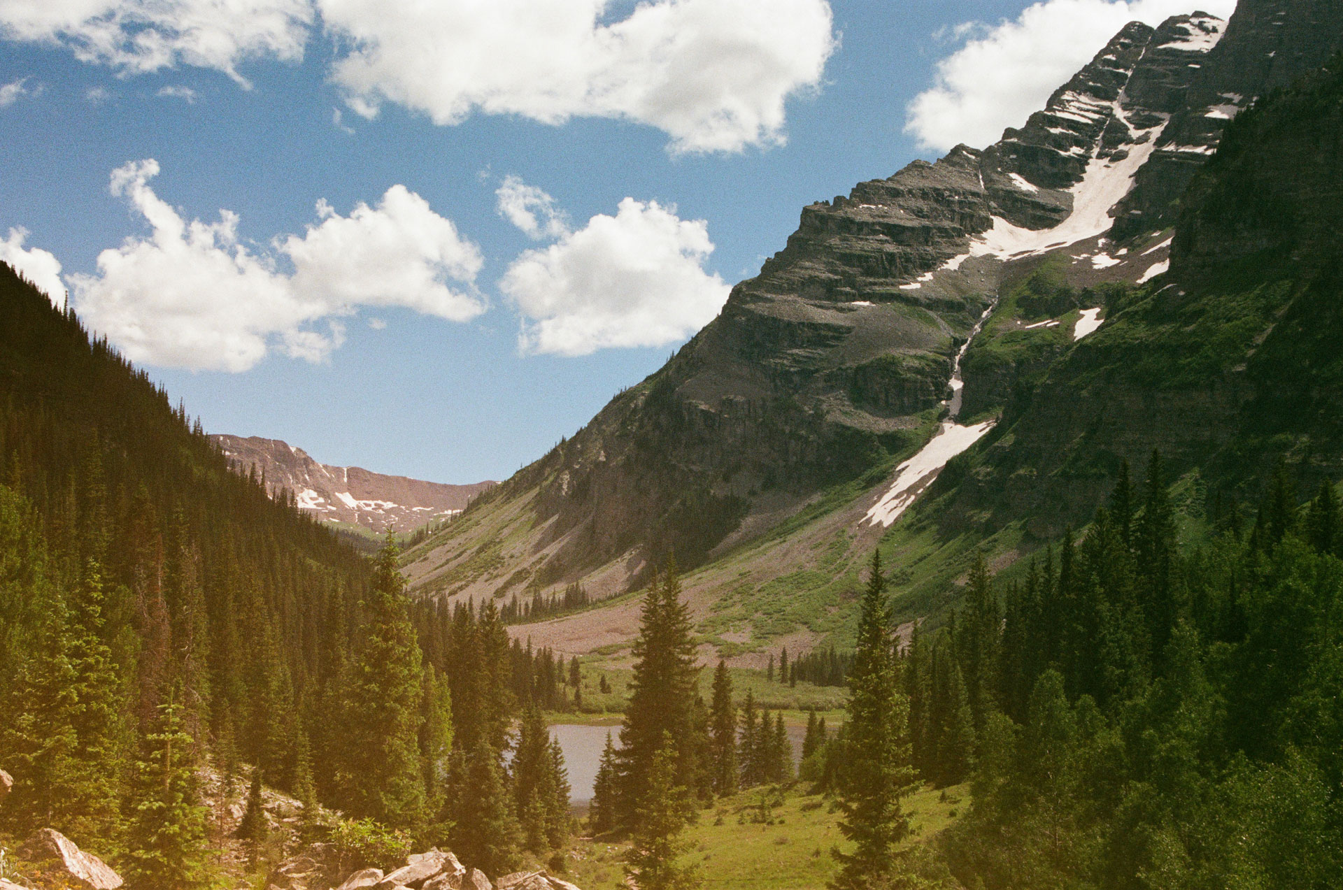 Mountain landscape near Aspen, Colorado, featuring rugged snow-dusted peaks, lush evergreen forests, and a small alpine lake nestled in the valley under a bright blue sky with scattered clouds.