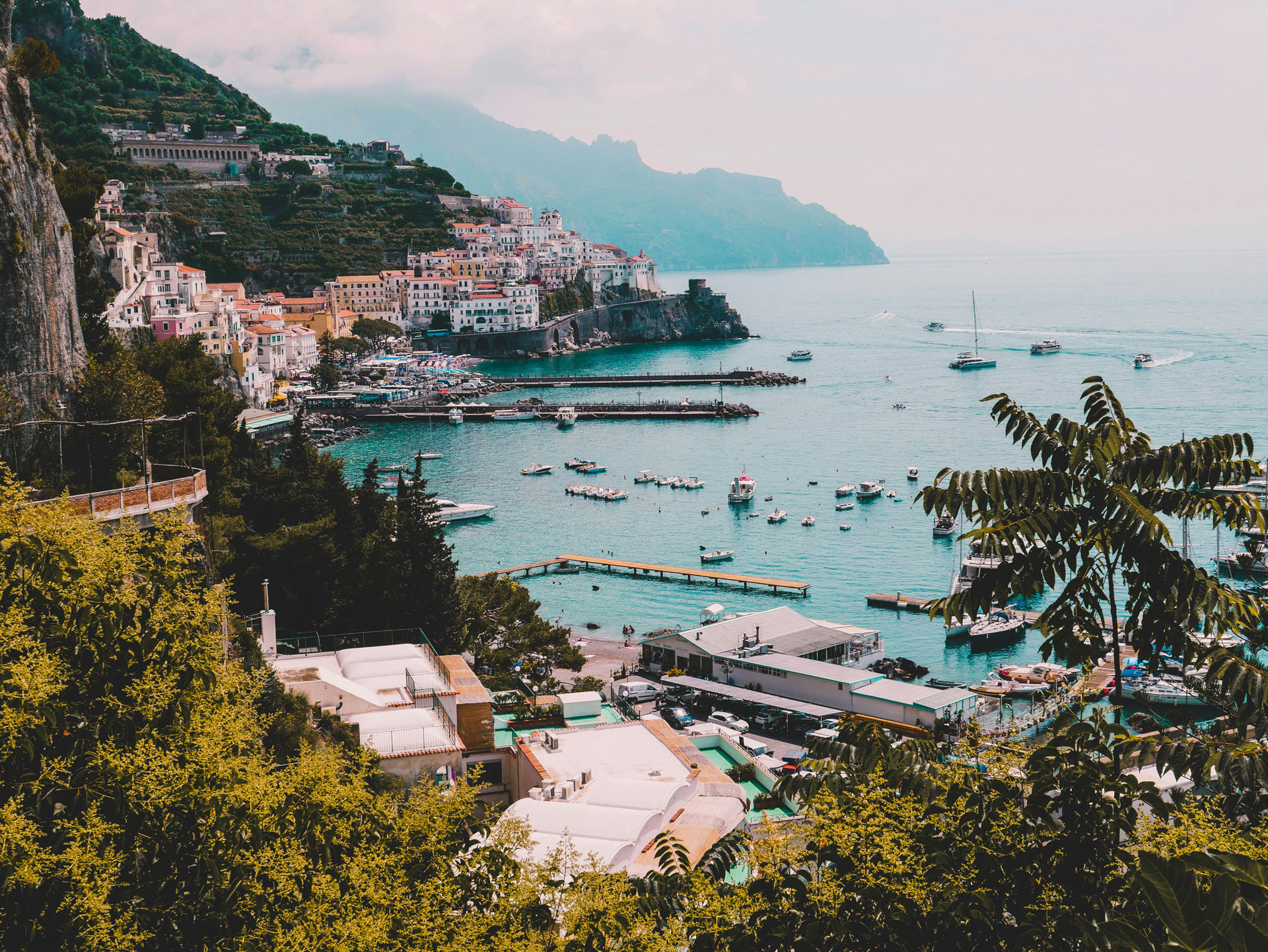 Picturesque view of the Amalfi Coast in Italy, featuring colorful cliffside buildings cascading down to a vibrant harbor filled with boats and yachts, set against turquoise waters and dramatic coastal mountains in the distance.