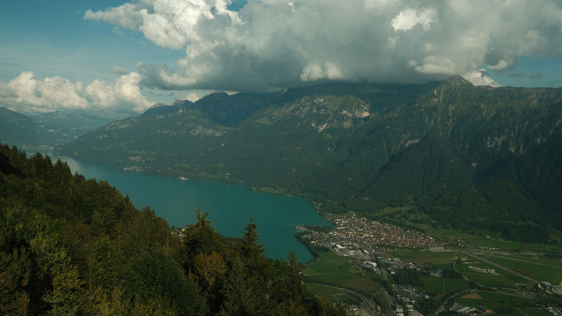 A sweeping view of Interlaken, Switzerland, nestled between lush alpine forests and turquoise Lake Thun, with dramatic mountain peaks rising in the background beneath a sky blanketed by thick, rolling clouds.